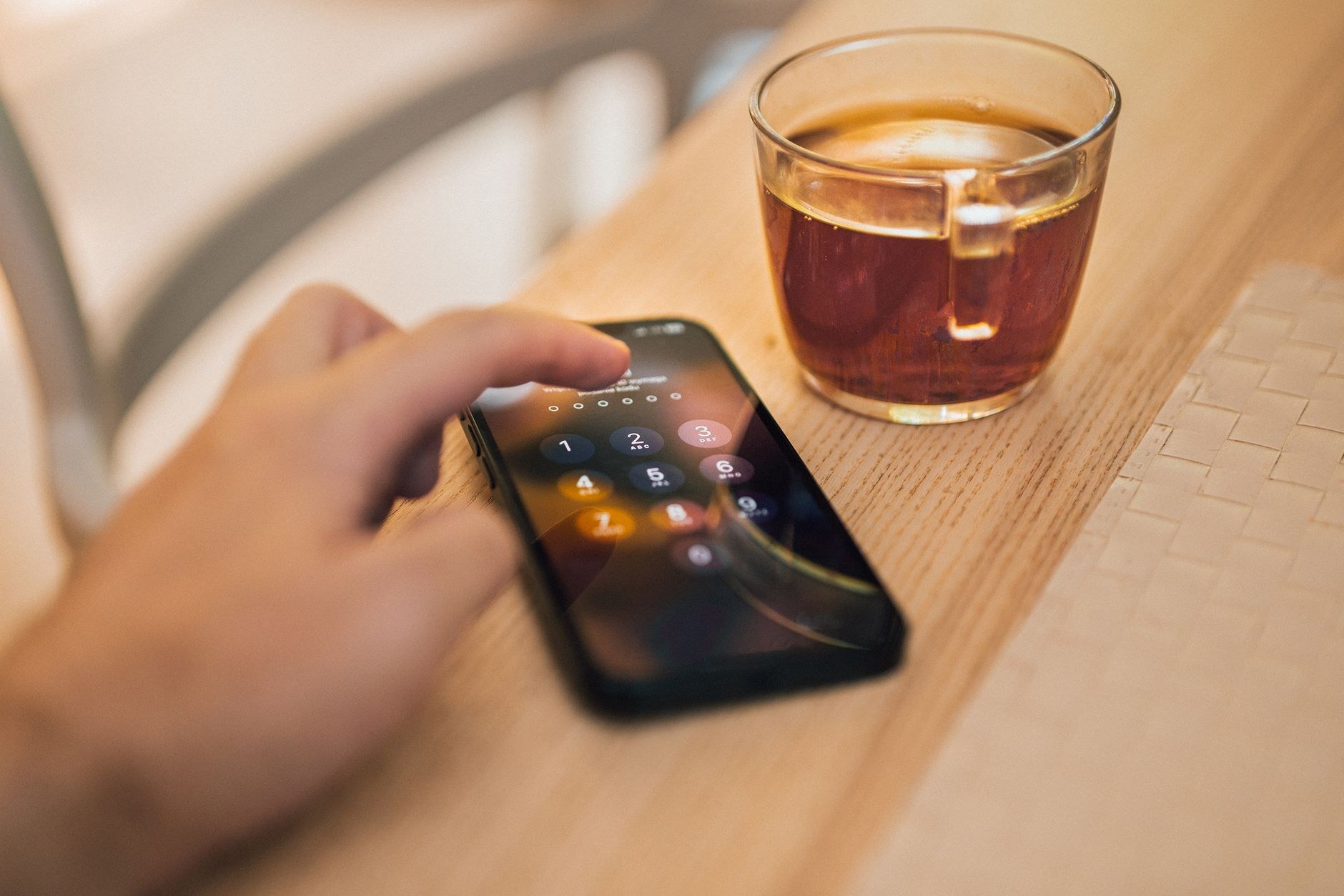 木のテーブルの上で、パスコード入力画面が表示されたスマートフォンに指で触れている様子。横にガラスのカップに入った紅茶が置かれている