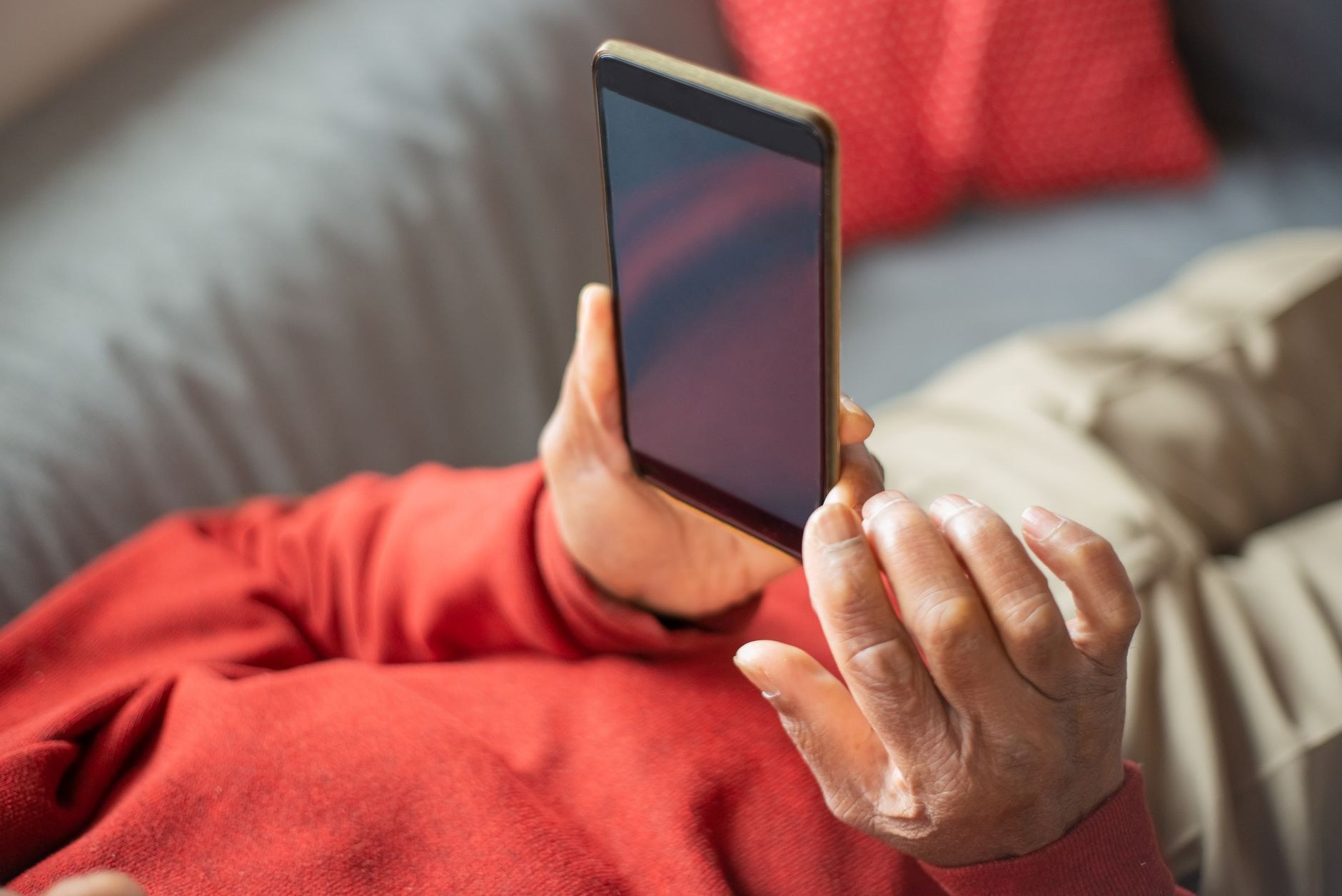 室内のソファに座った人が、画面に何も表示されていないスマートフォンを両手で持っている様子を手元に寄って写した写真