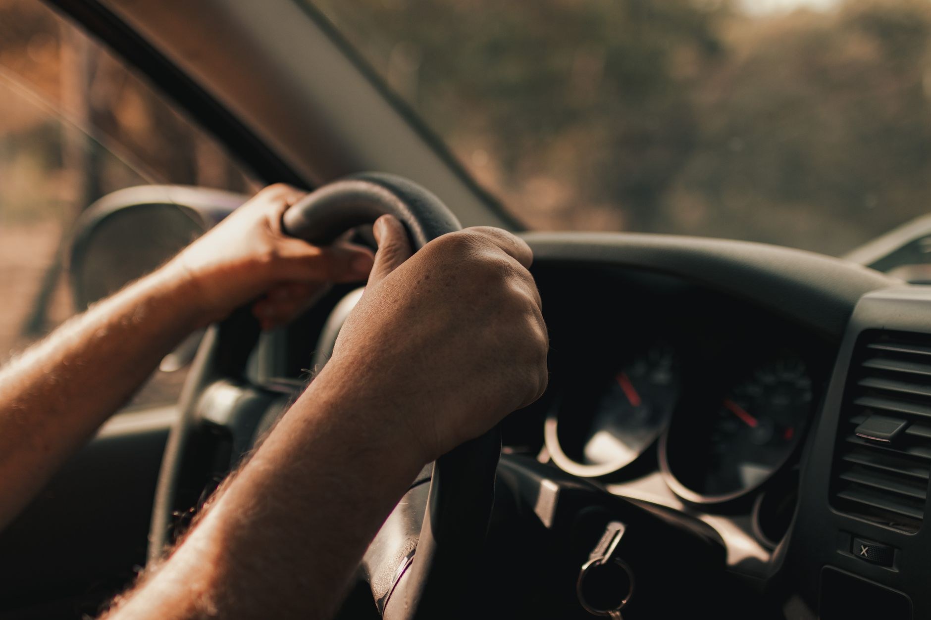 車のハンドルを両手で握っている運転席の手元を横から写した写真。メーターやダッシュボードがぼんやり写り、車種が分かるロゴや文字は入っていない